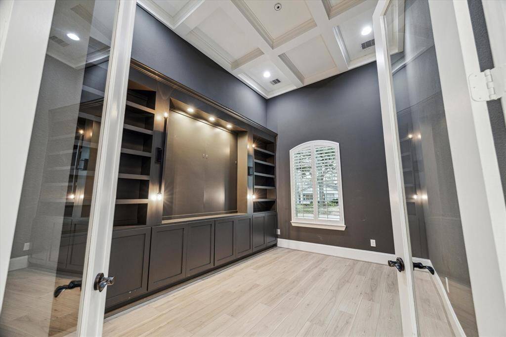 This room features a modern built-in shelving unit with accent lighting, a coffered ceiling, and large windows with shutters, offering a bright and stylish space perfect for a home office or library.