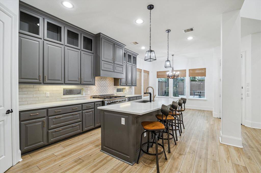 This modern kitchen features sleek gray cabinetry, a large island with seating, and stylish pendant lighting. The space is bright with natural light from large windows, and it has stainless steel appliances and vinyl flooring, offering a blend of elegance and functionality.