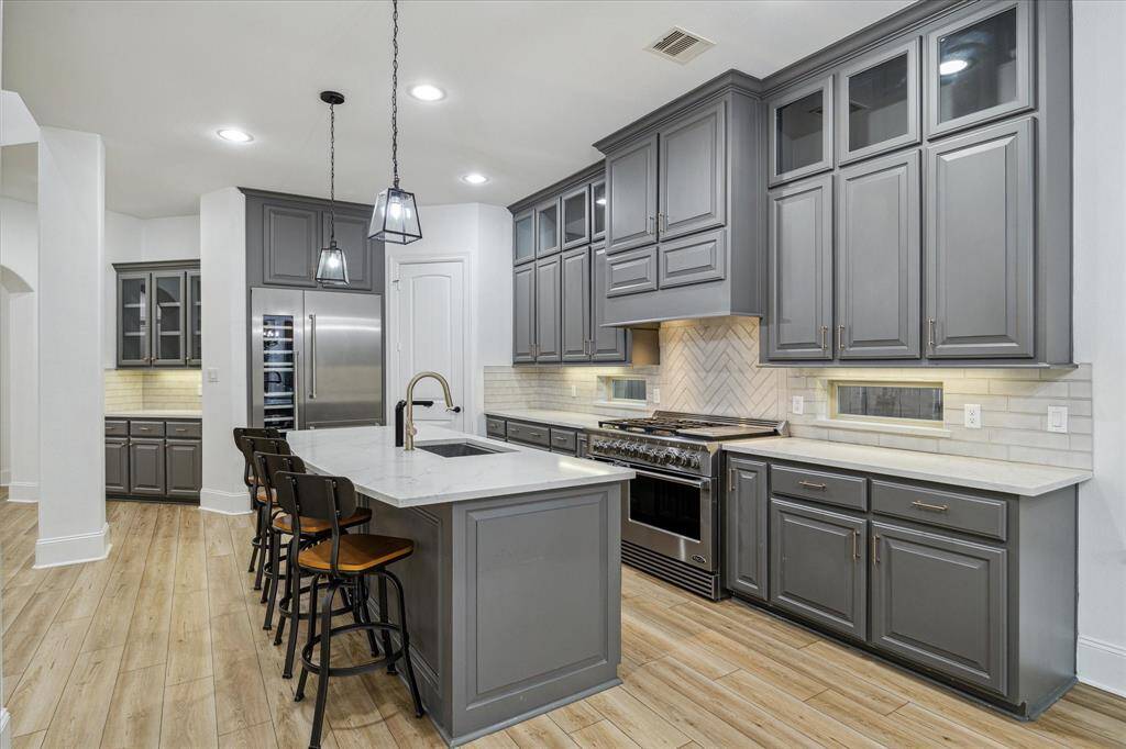 This modern kitchen features sleek gray cabinetry, stainless steel appliances, and a large island with seating. The herringbone backsplash adds a stylish touch, complemented by pendant lighting and vinyl flooring. Ideal for entertaining and everyday cooking.