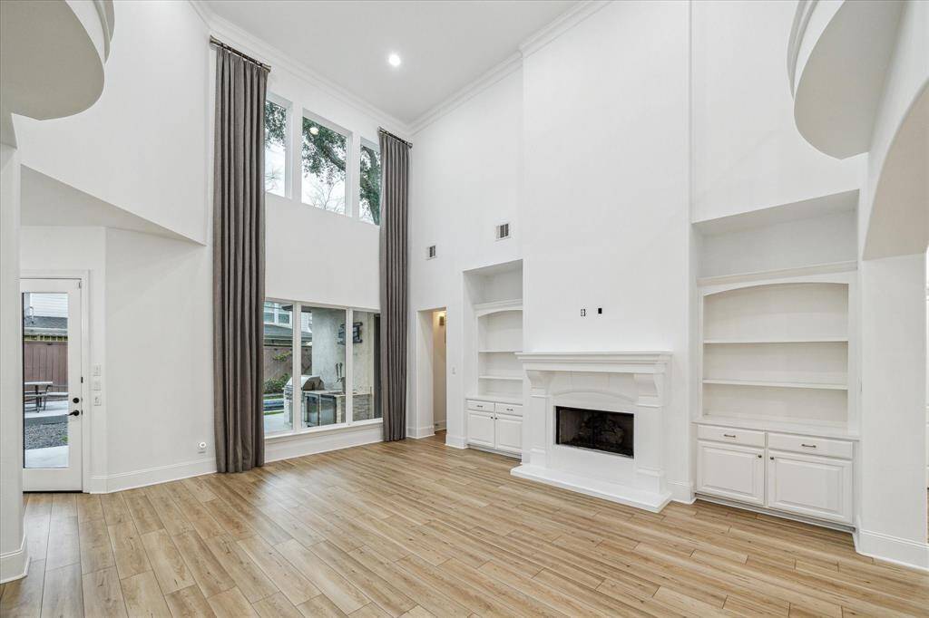This spacious living room features high ceilings, large windows with elegant drapes, and a cozy fireplace flanked by built-in shelves. The light vinyl flooring adds warmth, while a glass door provides access to the outdoor area.