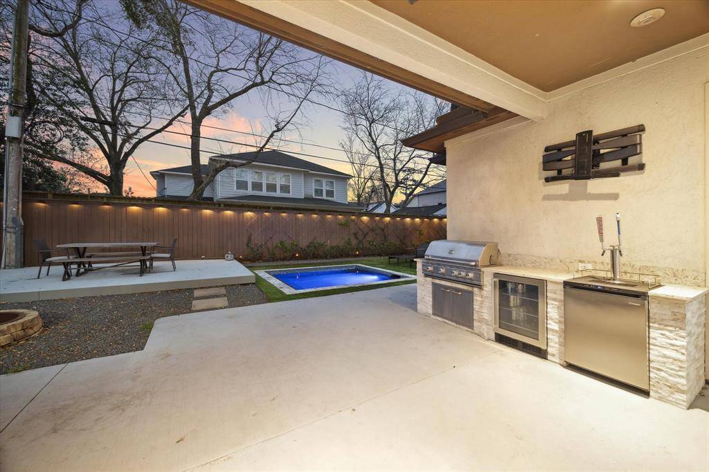 This photo features a modern backyard with a covered patio area, complete with an outdoor kitchen equipped with a grill, sink, and fridge. There's a cozy seating area nearby, a small pool/hot tub, and ambient lighting, creating an inviting space for relaxation and entertainment.