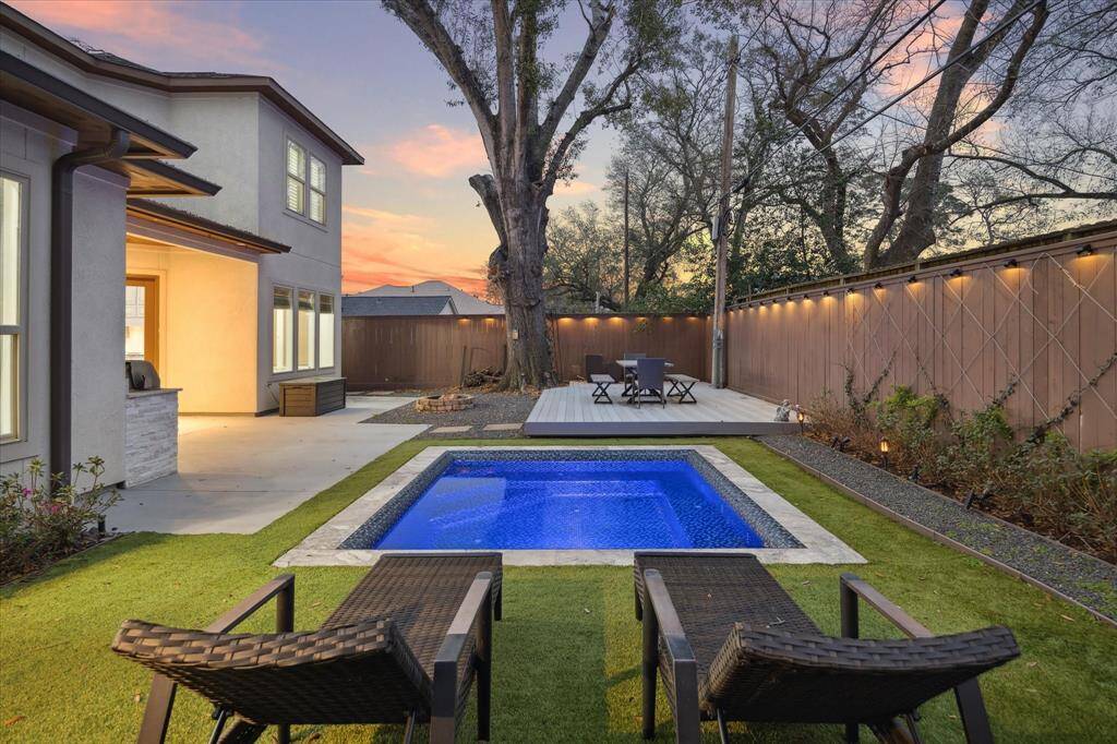 This backyard features a modern pool with surrounding greenery, a cozy patio area with seating, and ambient lighting. The contemporary home exterior complements the tranquil outdoor space, ideal for relaxation and entertaining.