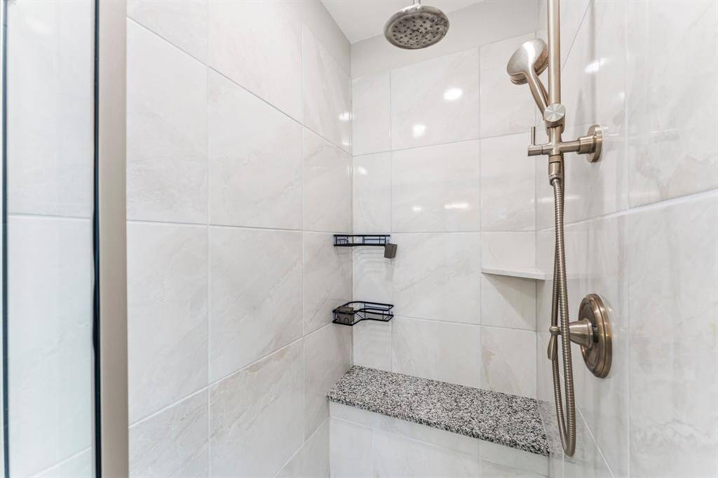 This photo shows a modern shower with elegant white tiles, a sleek rainfall showerhead, and a handheld sprayer. It features a granite bench and built-in shelving for convenience.