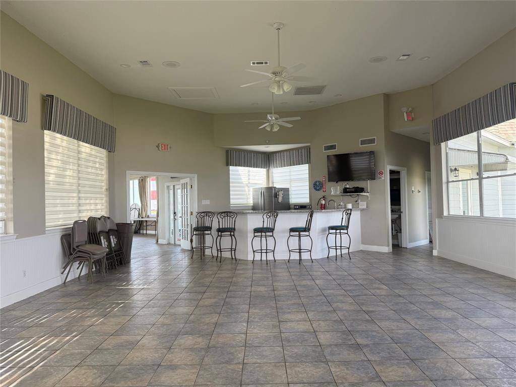 Spacious community room with a high ceiling and tiled floor. Features a kitchenette with a bar, stools, and stainless steel appliances. Large windows provide ample natural light, and there's a wall-mounted TV for entertainment. Ideal for gatherings and events.