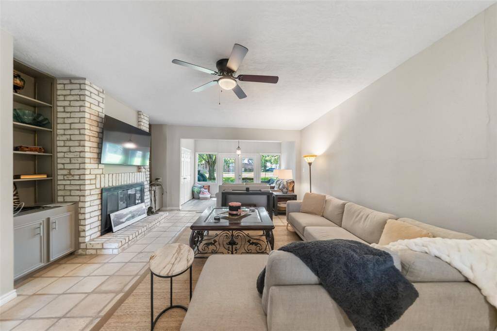 This spacious living room features a brick fireplace with a mounted TV. Natural light fills the room through large windows, and a ceiling fan adds comfort. The neutral tones create a warm, inviting atmosphere.
