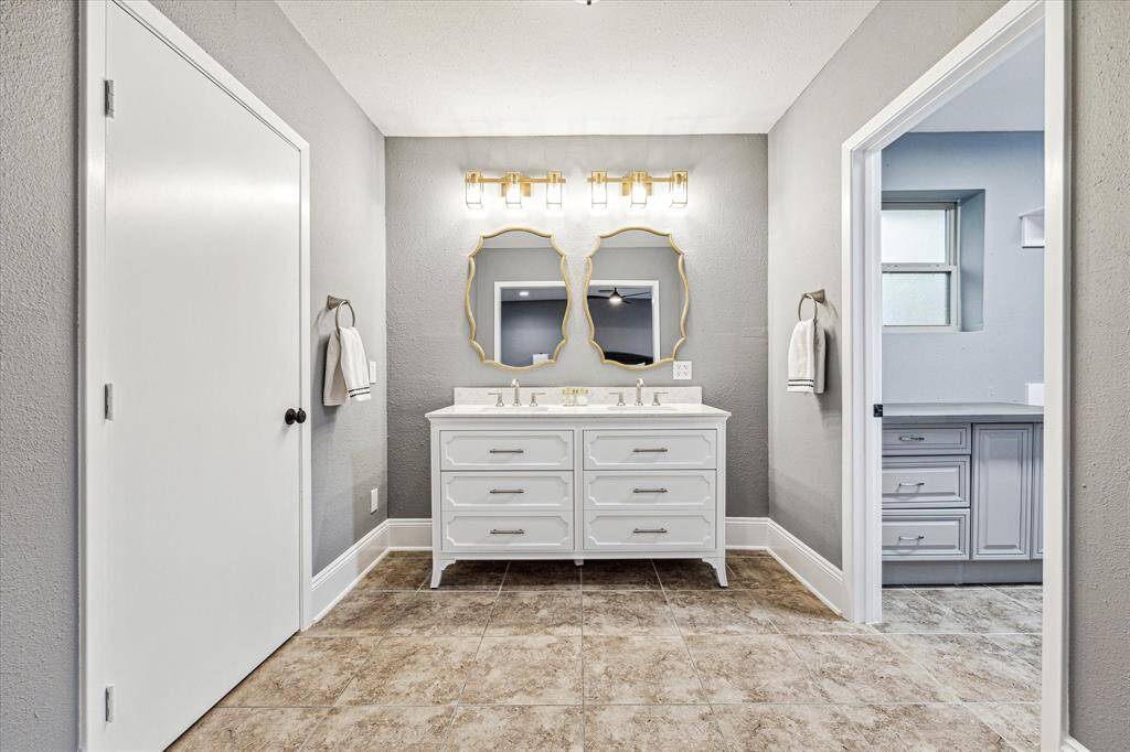 This primary suite offers a beautifully updated ensuite bathroom featuring a furniture style vanity with dual sinks with coordinating mirrors and vanity lights above. A massive walk-in closet with built-ins is to the right.