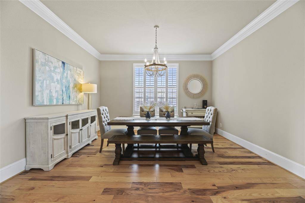 This dining room features elegant hardwood floors, a stylish chandelier, and a large window with shutters. It includes a wooden dining table with bench seating, a sideboard, and tasteful decor. Bright and welcoming, it's perfect for gatherings.