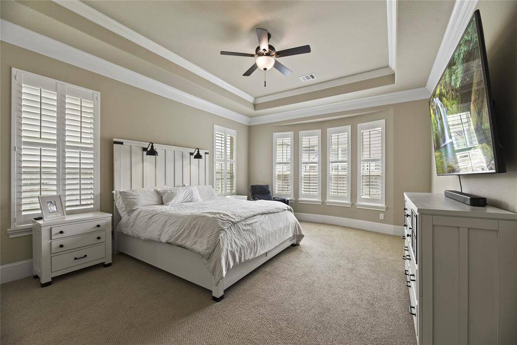 This bedroom features a spacious layout with a tray ceiling and ceiling fan. It has large windows with plantation shutters, allowing ample natural light. There’s a comfortable bed with wall-mounted lamps, a dresser, and a wall-mounted TV for entertainment. The neutral color palette enhances the room's serene ambiance.