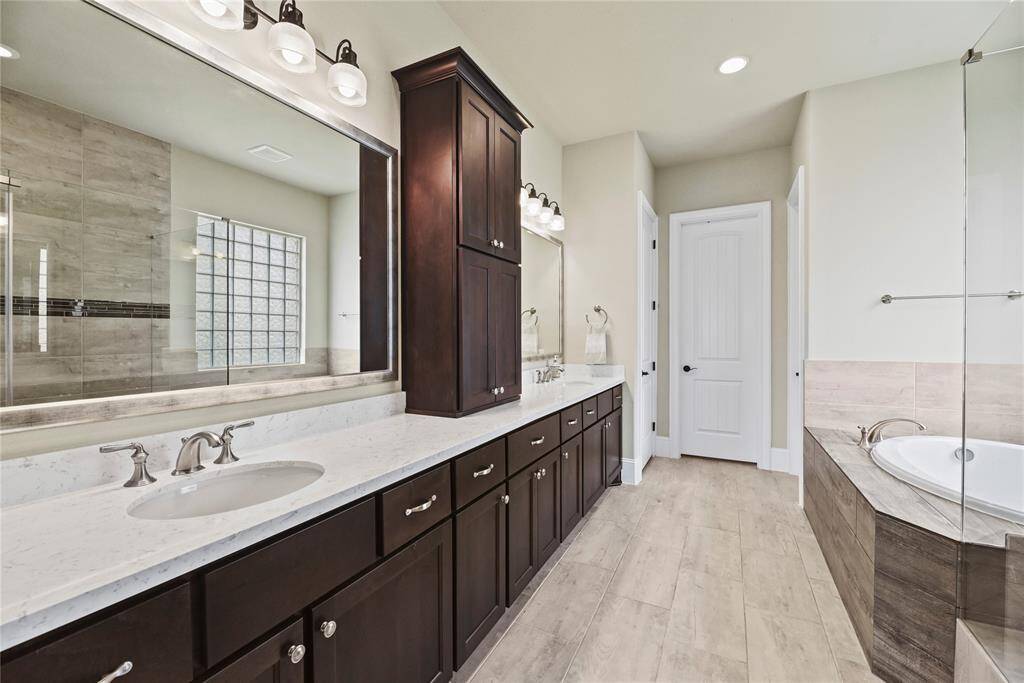 This bathroom features a spacious double vanity with dark wood cabinets, light countertops, and modern fixtures. It includes a large mirror, a walk-in shower with glass block accents, and a separate bathtub. The neutral tile flooring and soft lighting create a warm, inviting atmosphere.