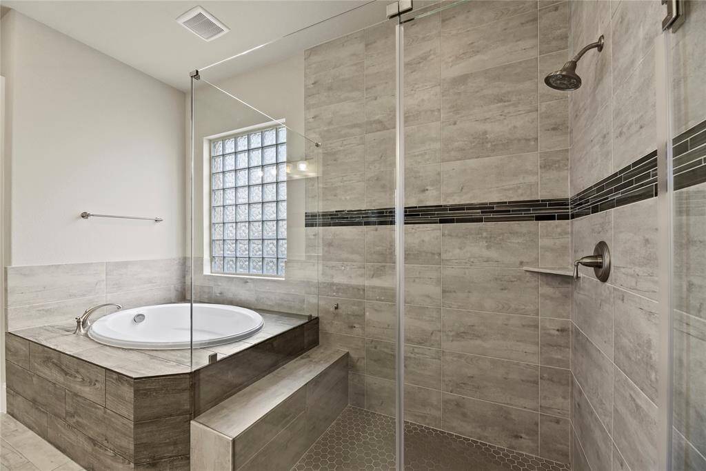 This bathroom features a modern design with a spacious walk-in shower and a corner bathtub. The neutral-toned tiles and glass block window enhance natural light, creating a bright and inviting atmosphere.