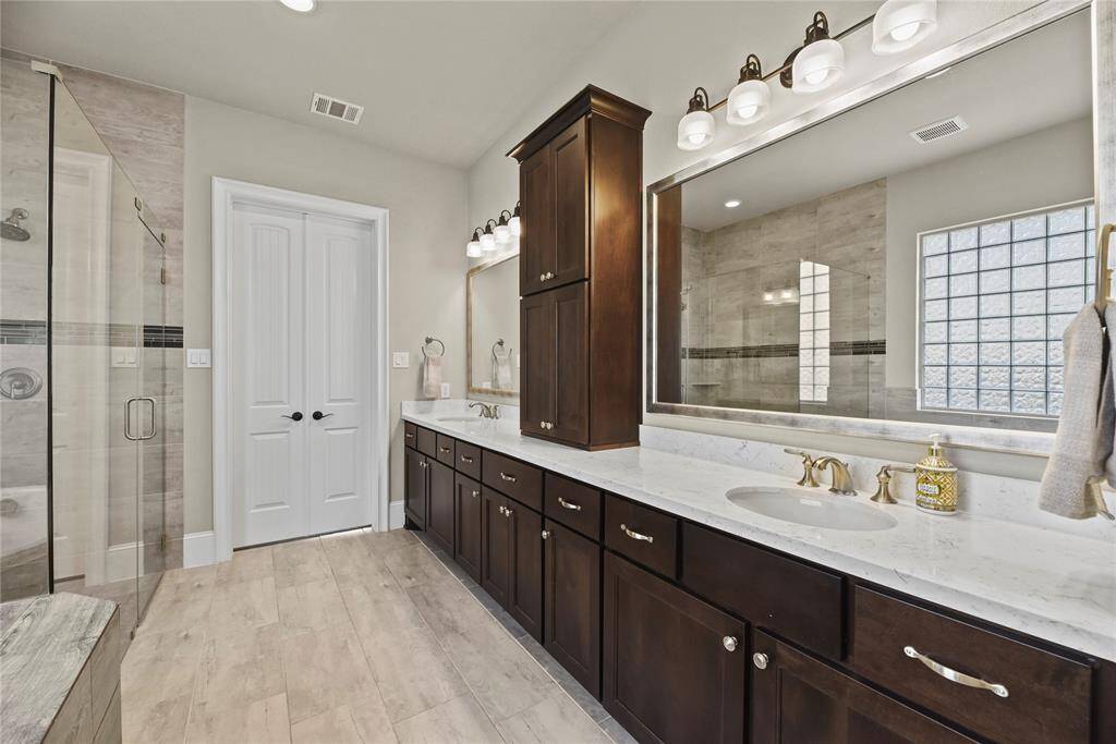 This bathroom features a spacious double vanity with dark wood cabinets, a large mirror, and elegant light fixtures. It includes a glass-enclosed shower with modern tile work and ample natural light from a glass block window, creating a bright and luxurious atmosphere.