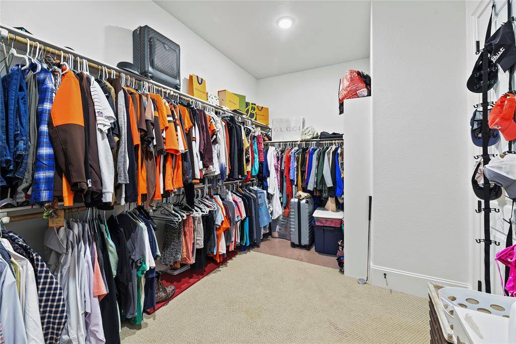 This photo shows a spacious walk-in closet with ample hanging space for clothes, organized shelves, and overhead storage. It's well-lit with a neutral carpeted floor, providing a functional and organized area for wardrobe needs.