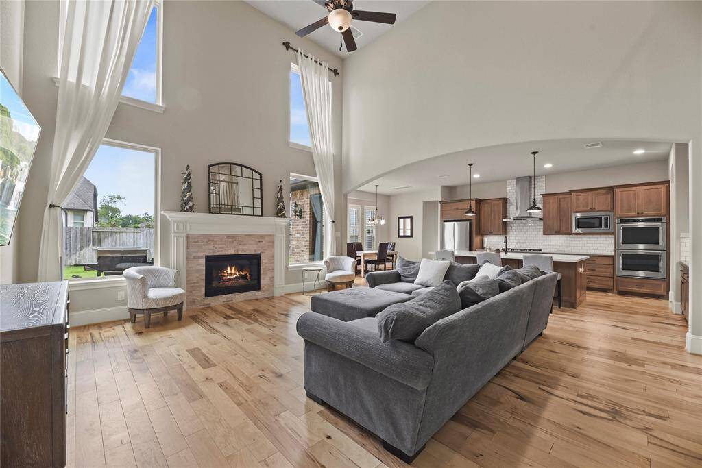 This spacious living area features high ceilings, abundant natural light, and a cozy fireplace. The open floor plan seamlessly connects to a modern kitchen with stainless steel appliances and warm wood cabinetry, perfect for entertaining.