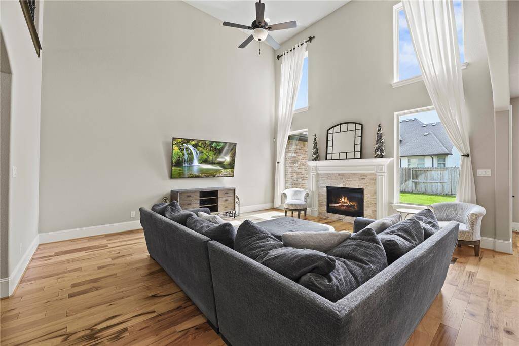 This spacious living room features high ceilings, abundant natural light from large windows, and a cozy fireplace. It includes a modern TV setup, a plush sectional sofa, and elegant hardwood floors. Perfect for relaxation and entertaining.