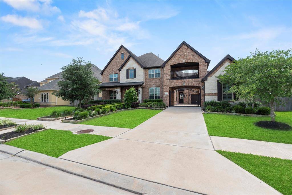 This photo showcases a spacious two-story brick home with a neatly manicured lawn, mature trees, and a long driveway leading to a gated entrance. The house features multiple roof gables, a covered balcony, and a welcoming front porch.