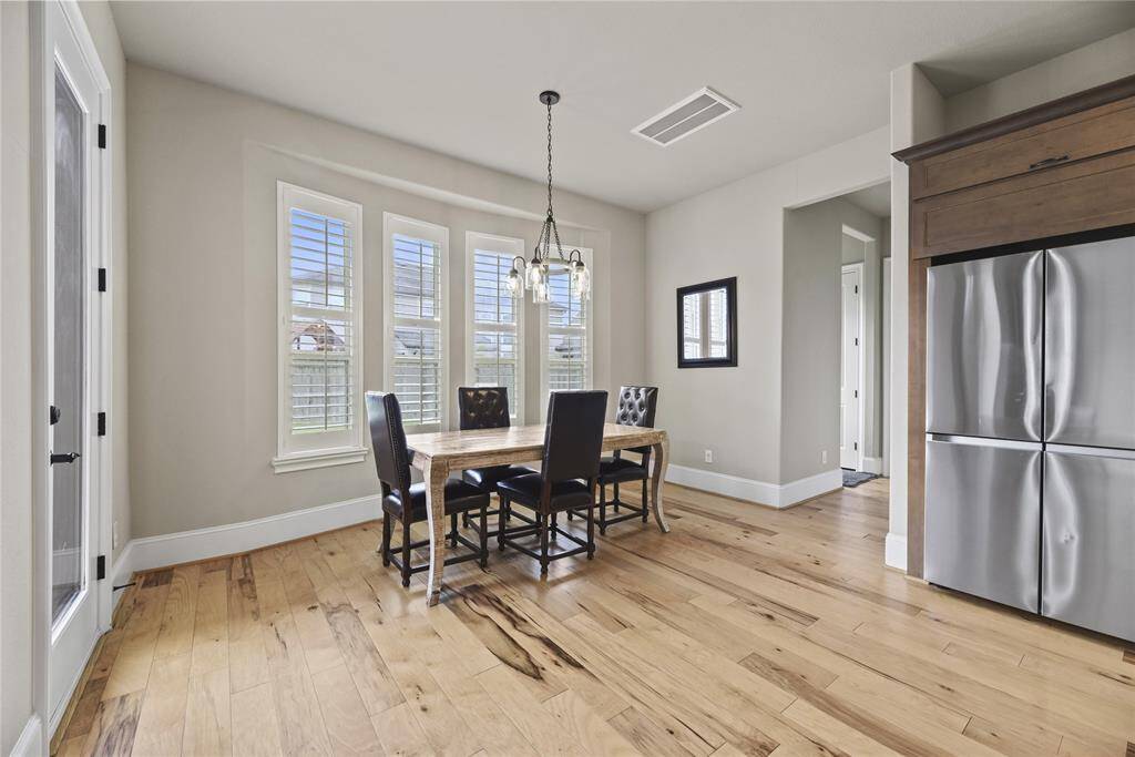 Bright dining area with large windows, hardwood floors, and a modern chandelier. Adjacent to a sleek stainless steel refrigerator, ideal for open concept living.