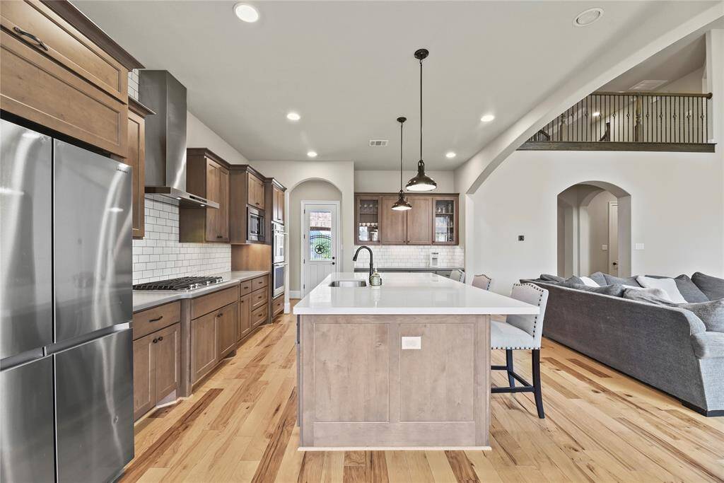 This photo showcases a modern, open-concept kitchen and living area. The kitchen features stainless steel appliances, a large island with seating, and wooden cabinetry. The space is bright with hardwood floors, pendant lighting, and a view of a cozy living room. An open loft area is visible above, adding to the airy feel.