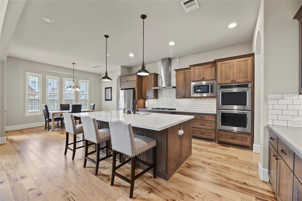 This spacious kitchen features modern stainless steel appliances, a large central island with seating, and elegant wood cabinetry. The open layout flows into a bright dining area with natural light from multiple windows, complemented by beautiful hardwood floors.