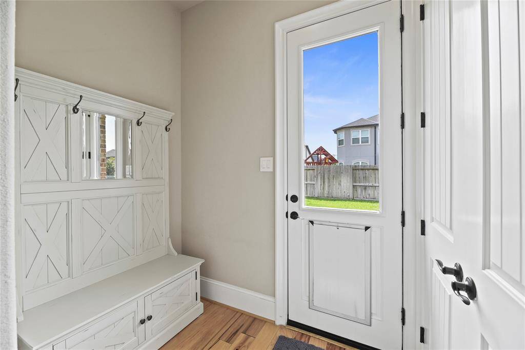 Bright and welcoming mudroom with a built-in bench and storage, featuring white paneling and coat hooks. A door with a window opens to a fenced backyard, offering a view of a neighboring house.