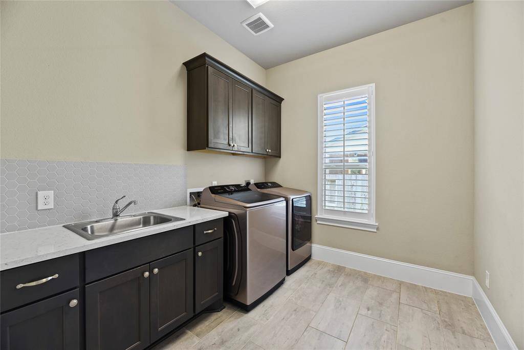 This photo showcases a modern laundry room with sleek appliances, a countertop with a sink, dark cabinetry, and a window with plantation shutters, offering a clean and functional space.
