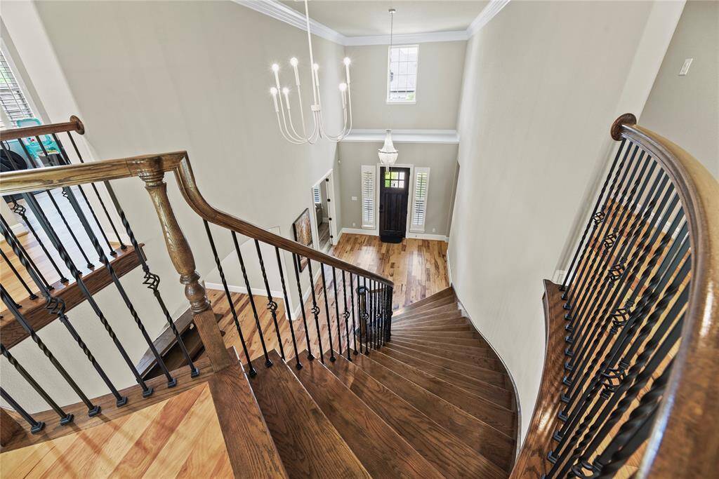 This photo showcases a grand, sweeping staircase with elegant wood and wrought iron railing, leading down to a bright, open foyer. The area features high ceilings, a modern chandelier, and a large, inviting front door, all complemented by polished wooden floors.
