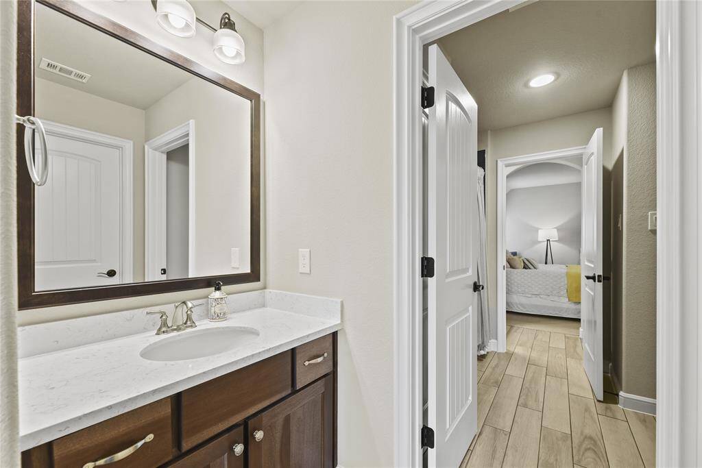 This photo showcases a modern bathroom with a sleek marble countertop and a large mirror, leading into a cozy bedroom with wood-style tile flooring and soft lighting.