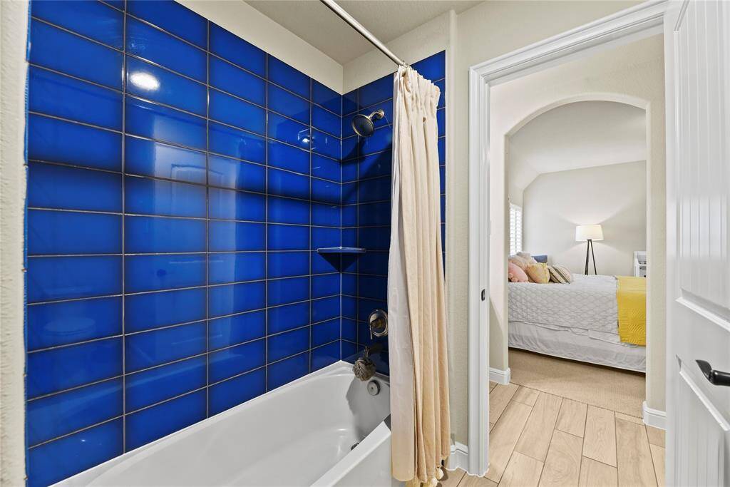 This photo shows a bathroom with vibrant blue tiles in the shower area and a curtain. It leads into a cozy bedroom with a modern lamp and neutral bedding, offering a warm and inviting atmosphere.