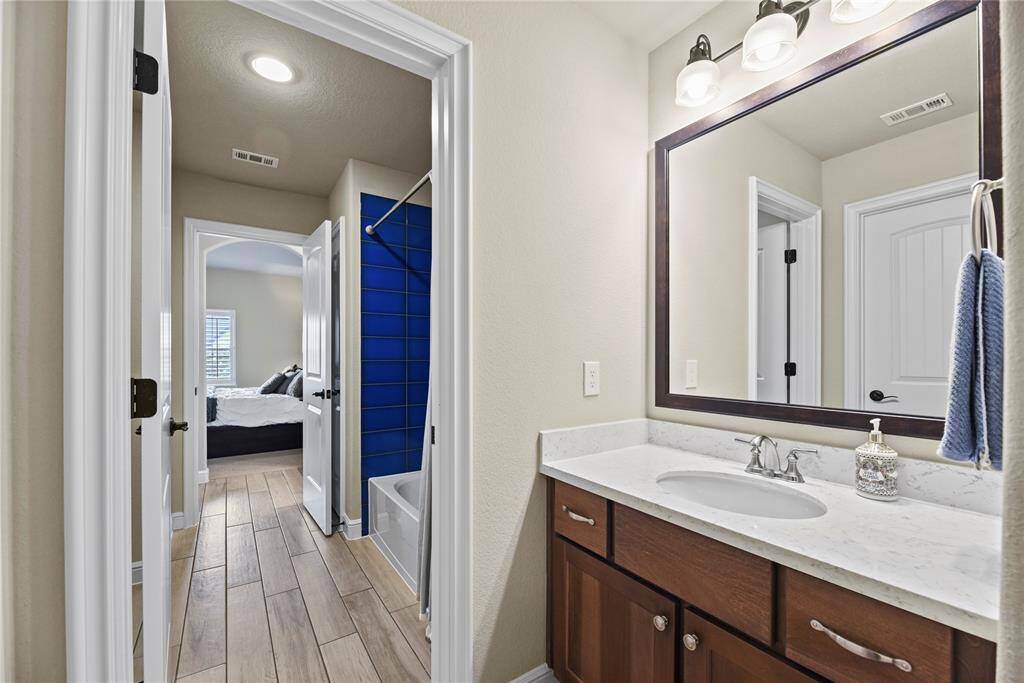 This photo shows a bright bathroom with a modern vanity featuring a large mirror and stylish lighting. The countertop is white, and there are rich wooden cabinets below. The room leads to a bedroom with natural light, visible through an open door. The bathroom also includes a shower with bold blue tiles.