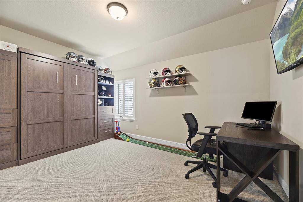 This is a versatile home office or hobby room featuring a built-in Murphy bed, a desk with a computer setup, ample shelving for display, and neutral carpeting. Ideal for working from home or as a multifunctional space.
