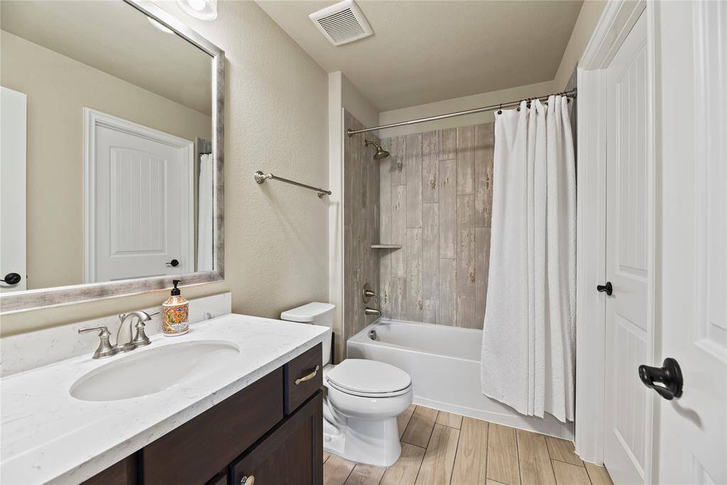 This bathroom features a modern design with a sleek vanity, marble countertop, and a large mirror. It includes a bathtub with a shower, surrounded by stylish wood-look tiles, and has neutral walls and flooring for a clean, inviting look.