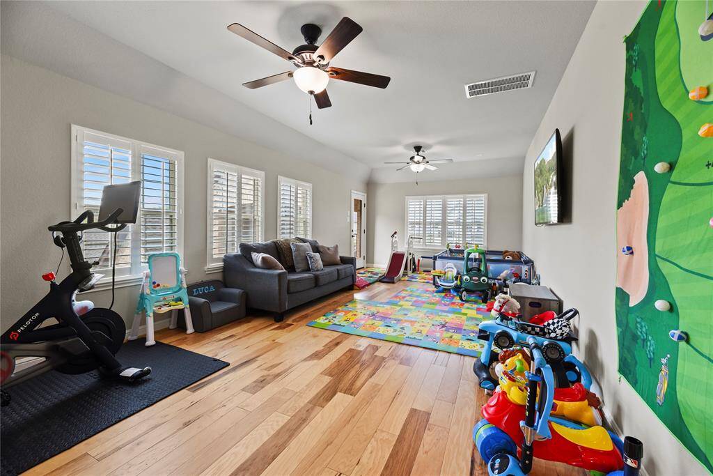 Bright, spacious playroom with hardwood floors, ceiling fans, and large windows. Features include a cozy sofa, exercise bike, colorful play mats, and ample space for toys and activities, making it ideal for a family.