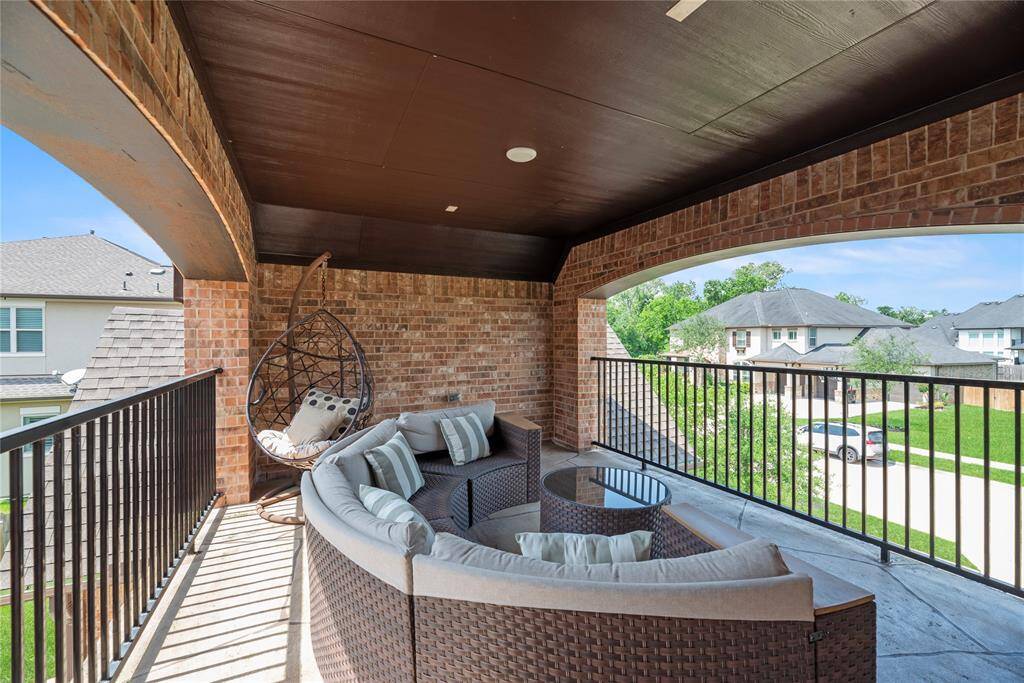 This photo showcases a spacious, covered balcony with brick accents, featuring comfortable outdoor seating and a hanging chair. It overlooks a quiet residential neighborhood, offering a relaxing space with scenic views.