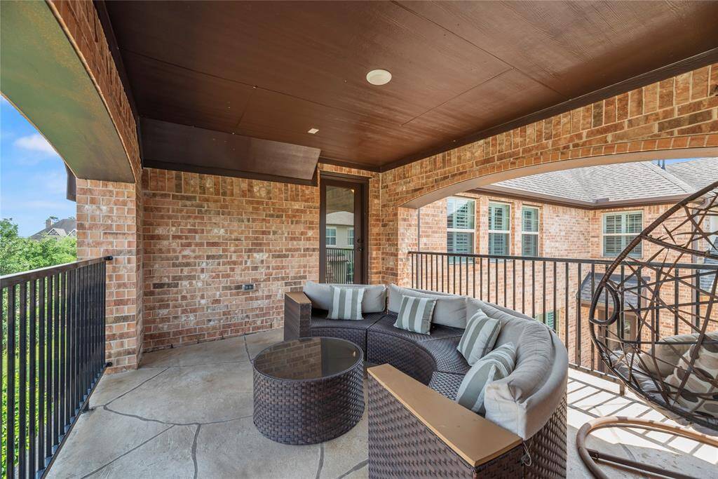 This inviting covered balcony features a cozy seating area with a sectional sofa and a hanging chair, surrounded by elegant brickwork and wrought iron railing. Perfect for relaxation and enjoying the view.