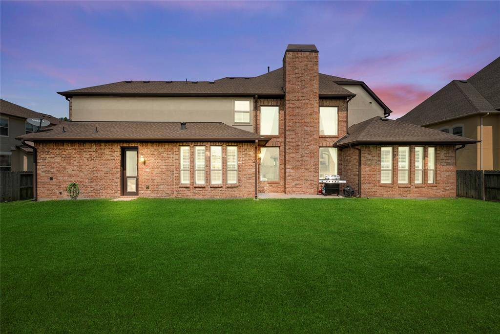 This photo showcases the rear view of a spacious two-story brick home with a large, well-maintained lawn, ample windows, and a central chimney. The setting sun casts a warm glow, highlighting the inviting backyard space.