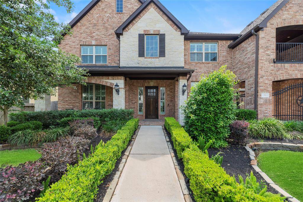 This charming two-story brick home features a beautifully landscaped front yard with lush greenery and a welcoming path leading to a classic wooden front door. The exterior combines brick and stone accents, offering a timeless and elegant appearance.