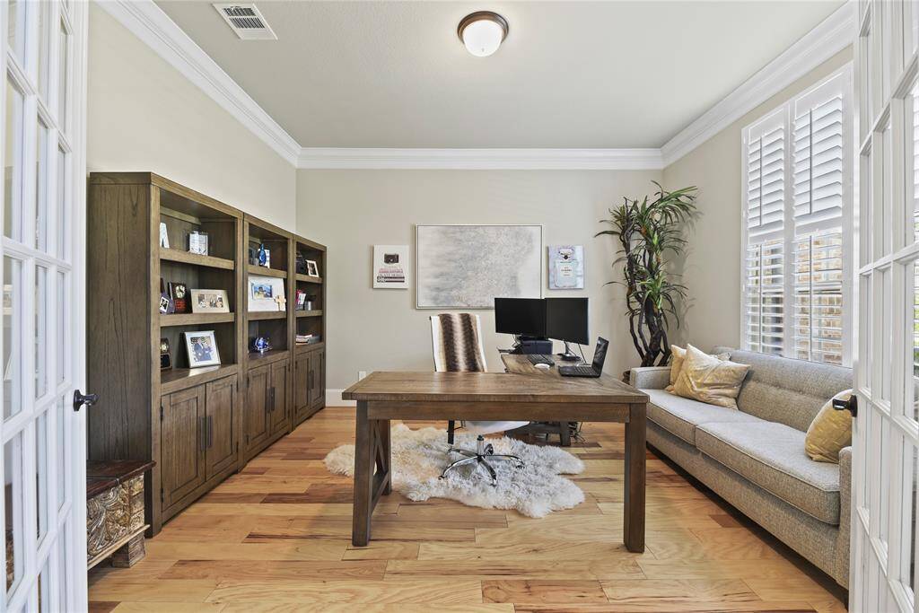 This inviting home office features a spacious wooden desk, built-in shelving, and ample natural light from large windows with shutters. The room is complemented by a cozy sofa, light wood flooring, and tasteful decor, creating a perfect workspace.
