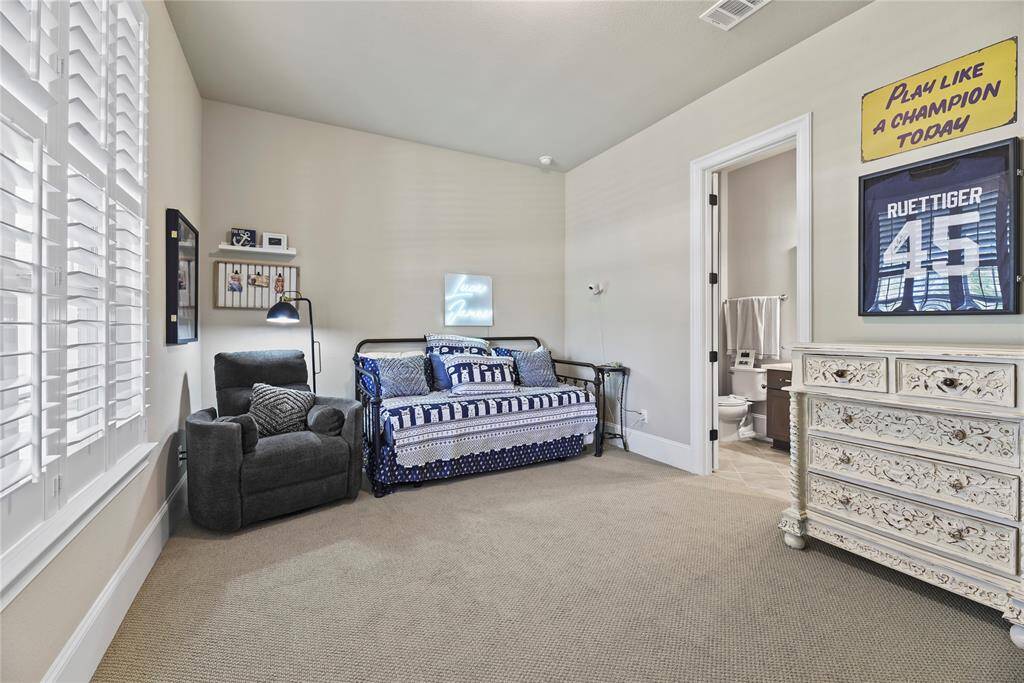 This cozy bedroom features carpeted flooring, a daybed with decorative pillows, and a comfortable armchair. It includes a framed sports jersey for a personal touch, plantation shutters on the window for natural light, and an adjoining bathroom for convenience.