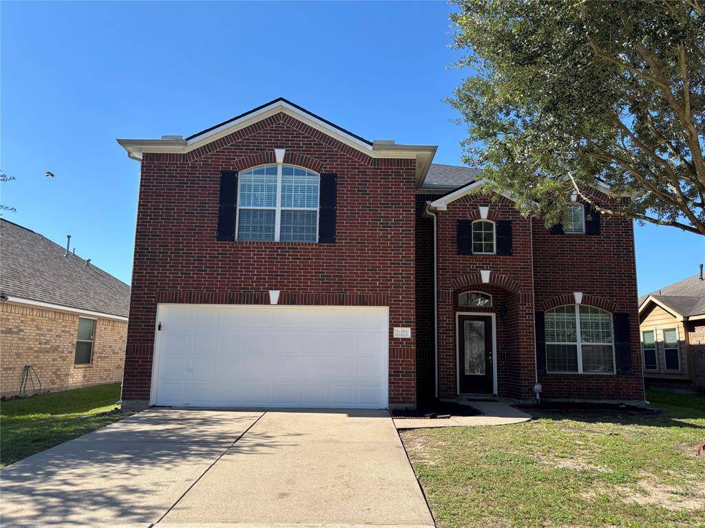 This photo shows a two-story brick home with a gabled roof and a large white garage door. It features a tidy front lawn and a driveway leading to the entrance, which is accented by arched windows and dark shutters.