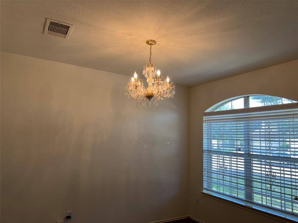 This is a formal living room which is on the right side as you enter the house. It features a classic chandelier and a large window with blinds, offering plenty of natural light. The ceiling is simple, and the walls are painted a neutral color, creating a versatile space for dining or other uses.