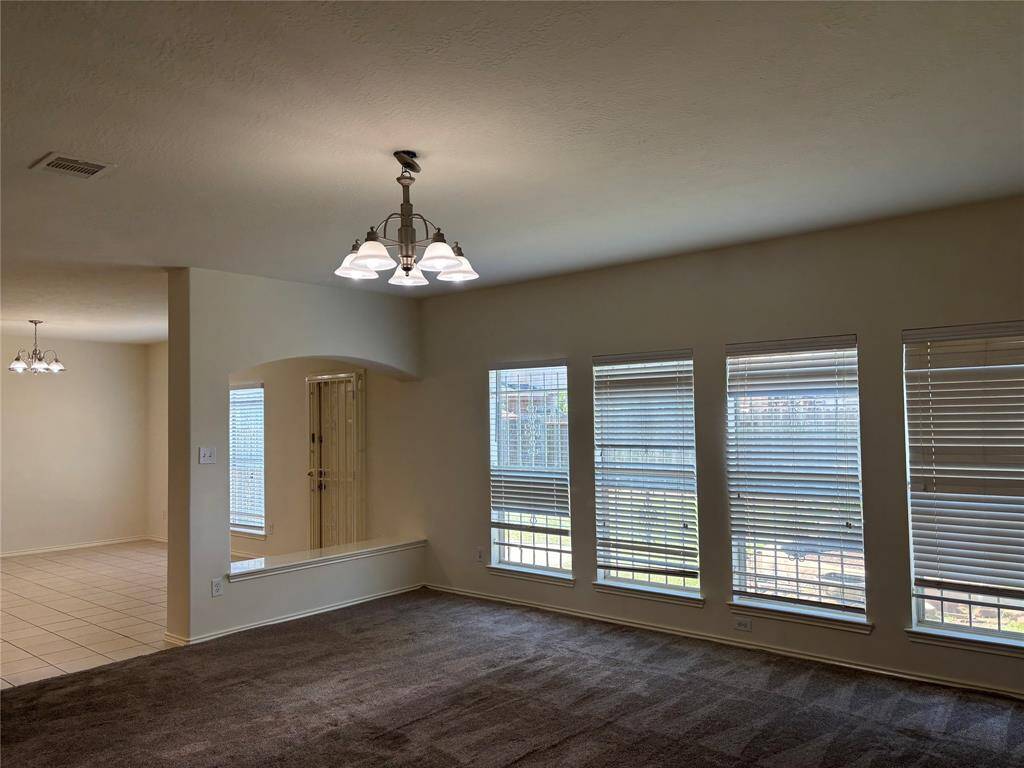 This spacious Family room features large windows with blinds, providing ample natural light. It has a carpeted floor and a stylish overhead light fixture. The open layout includes a view into a tiled dining area, enhancing the sense of space.