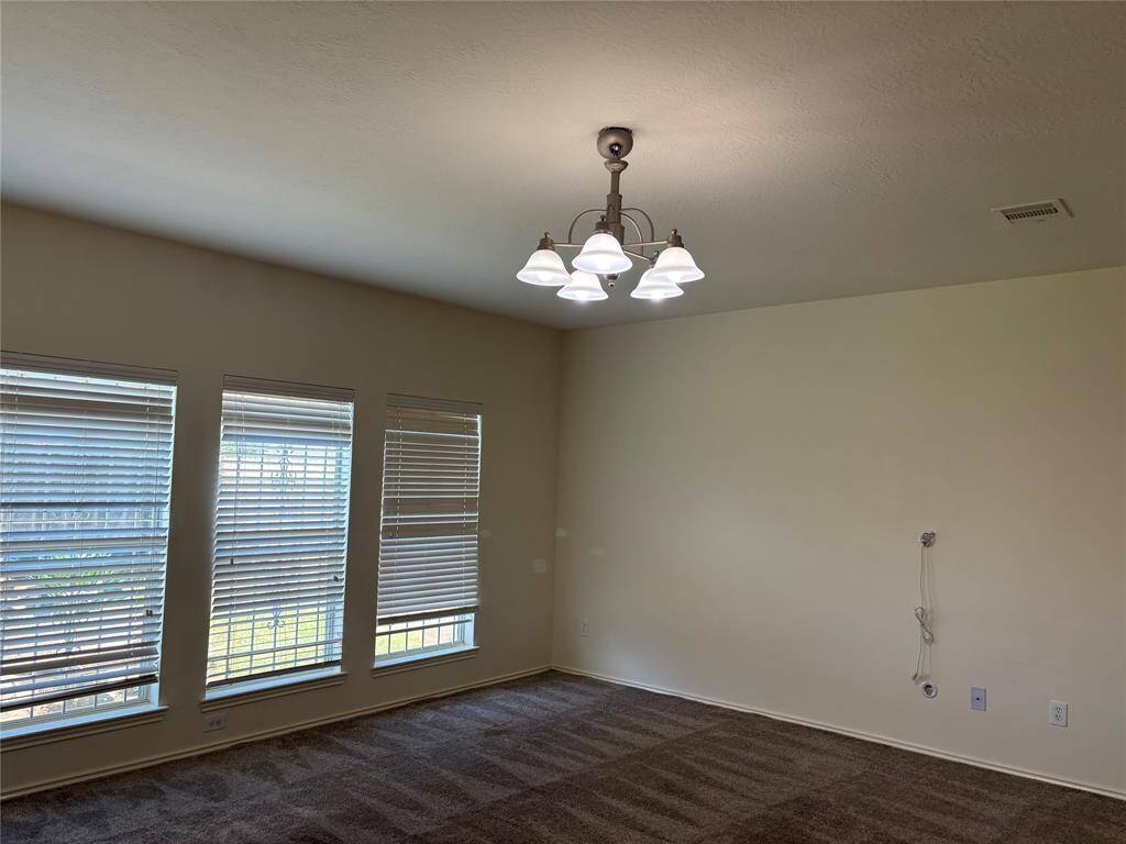 This family room area features a spacious area with carpeted flooring, large windows with blinds that allow natural light, and a ceiling light fixture. The walls are a neutral tone, offering a blank canvas for personalization.