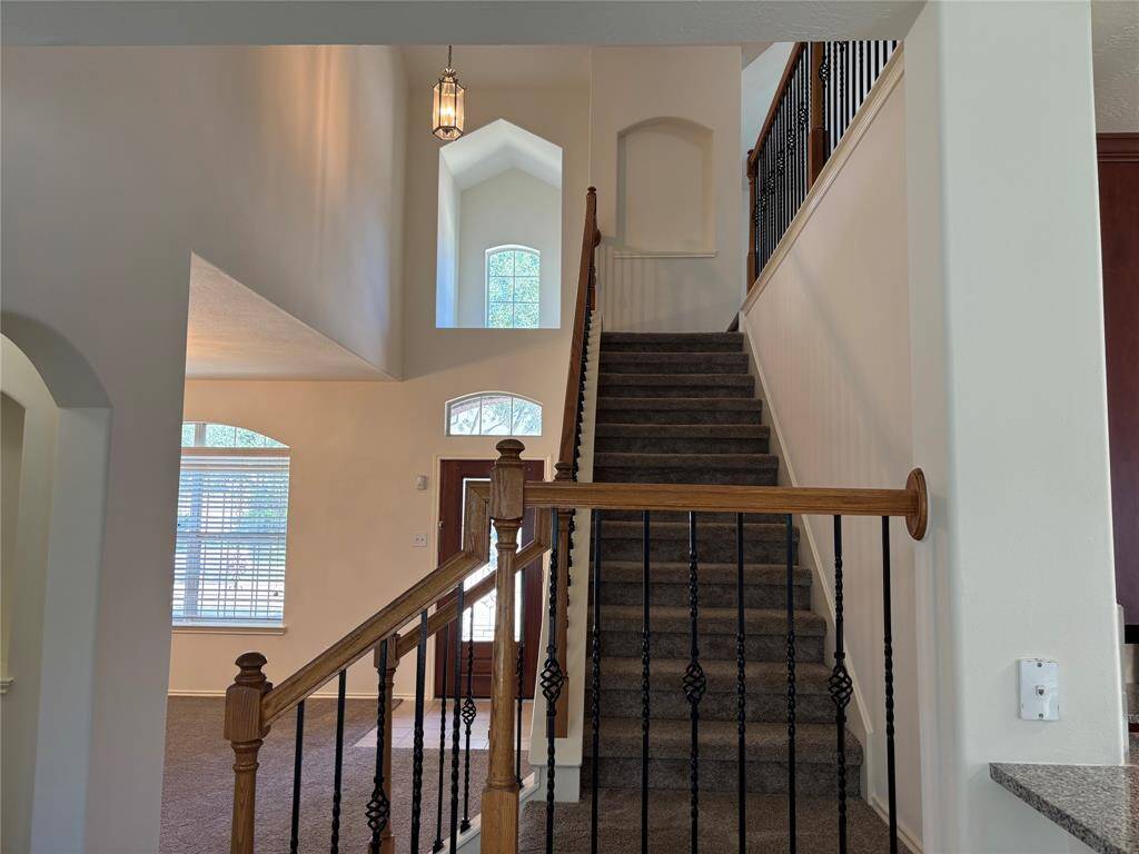 This photo showcases a bright entryway with a stylish staircase featuring wooden handrails and decorative iron balusters. The space is enhanced by high ceilings, arched windows, and an elegant light fixture, offering a welcoming and open atmosphere.