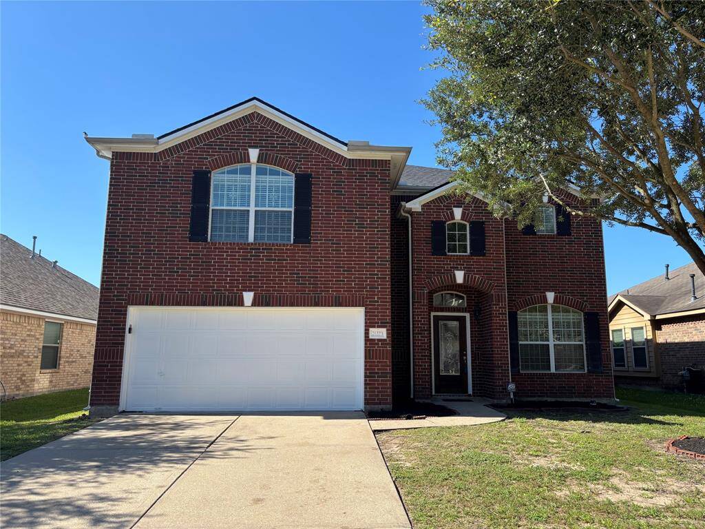 This is a two-story brick home featuring a two-car garage and arched windows. It has a neatly maintained front yard and a driveway, with a tree providing some shade on the right side.