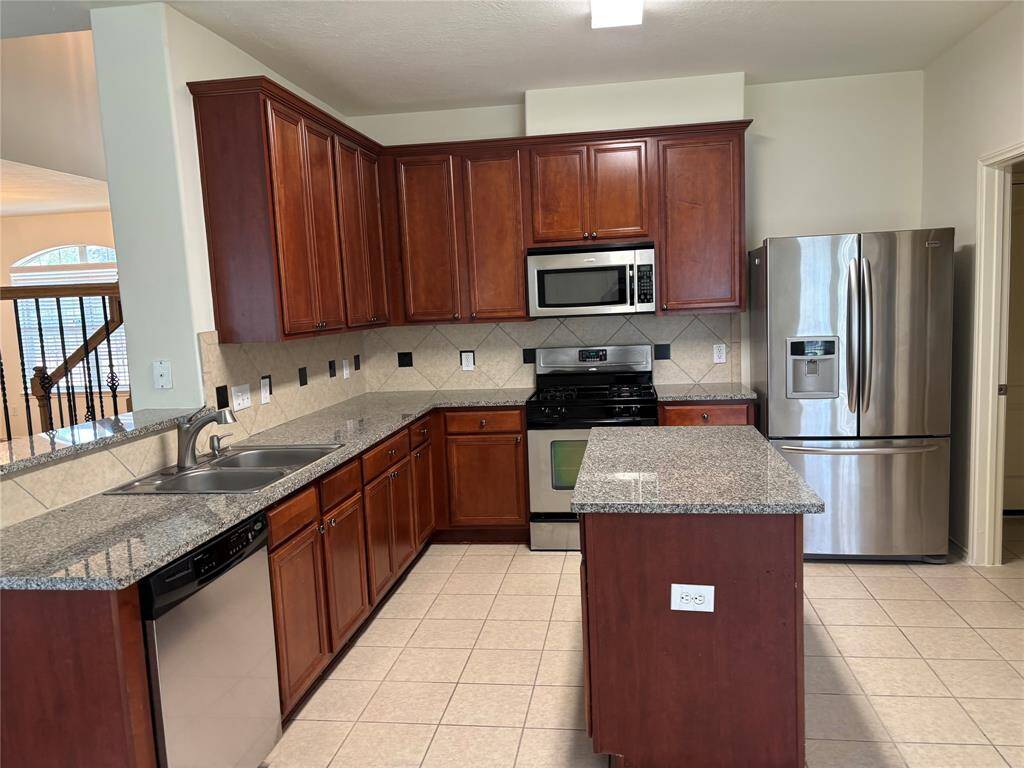 This modern kitchen features dark wood cabinets, granite countertops, and stainless steel appliances, including a fridge, stove, and dishwasher. It has a central island and tile flooring, providing a spacious and functional cooking area. An open entryway with stairs is visible in the background.