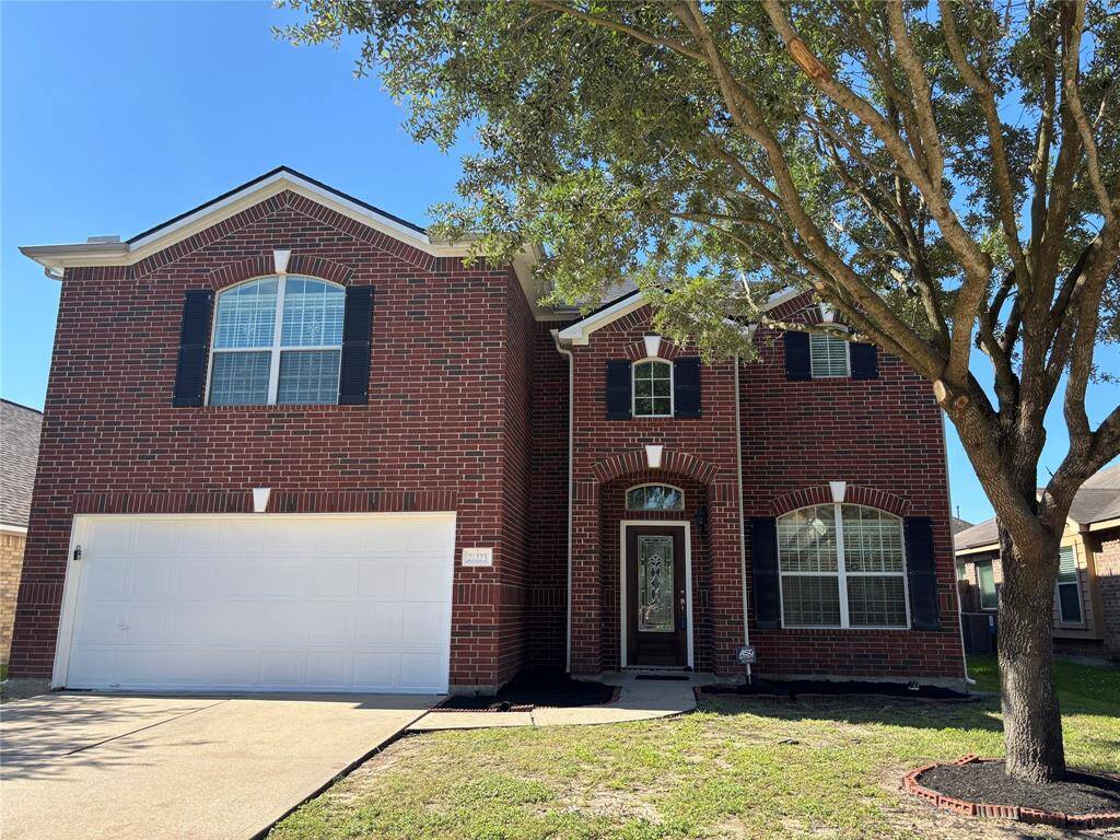This is a two-story brick home featuring a double garage and a welcoming front entrance. A large tree provides shade to the front yard, adding curb appeal.