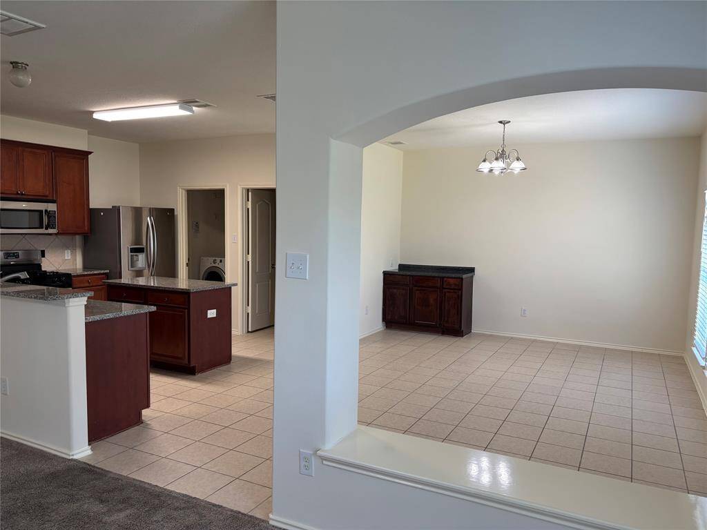 This photo showcases an open-concept space with a tiled floor and neutral walls, featuring a kitchen area with granite countertops and stainless steel appliances. Adjacent is a dining area with a chandelier, a window providing natural light, and a convenient laundry room nearby.