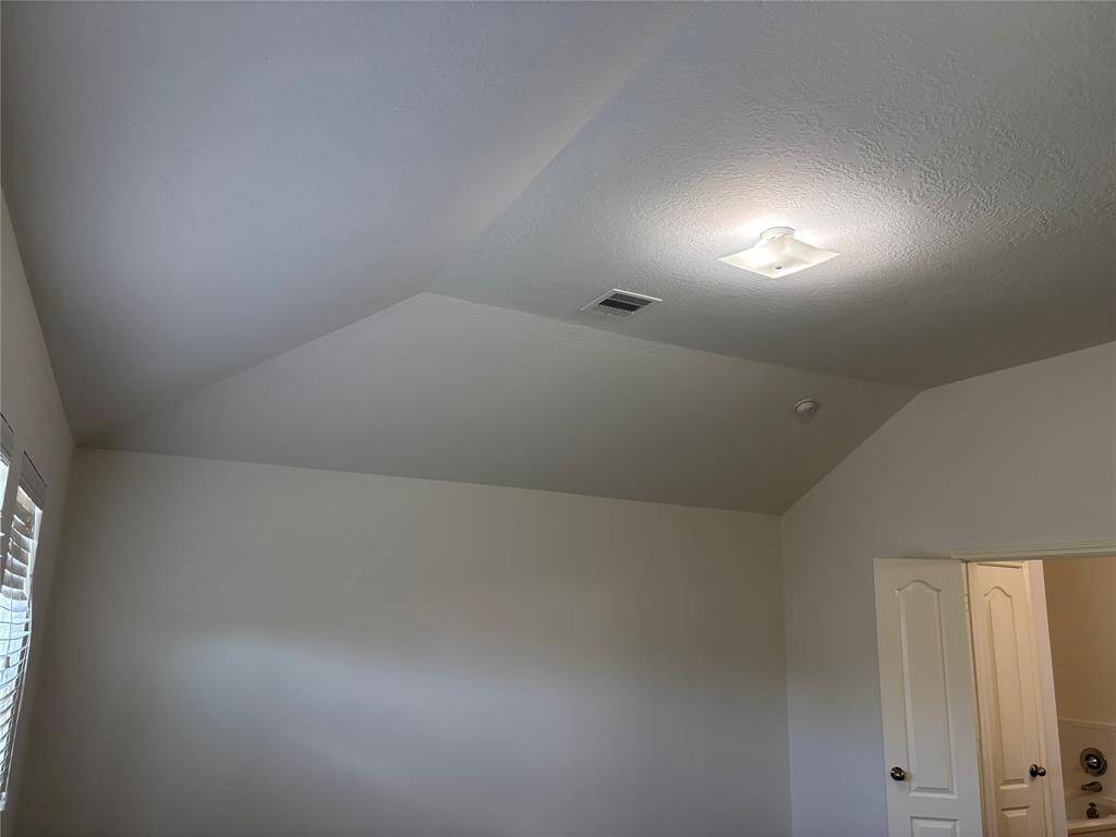 This photo depicts a small, neutral-color room with carpeted flooring. It features a window with blinds allowing natural light in, and a ceiling light fixture.