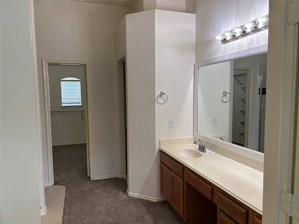 This photo showcases a bathroom with a spacious countertop, a large mirror, and wooden cabinets. It leads to a carpeted walk-in closet with a window, providing ample natural light.