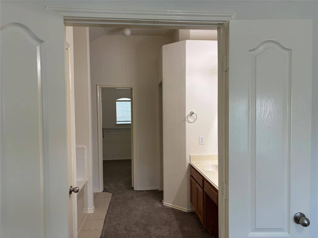 This photo shows a bathroom with double doors leading to a vanity with a countertop and sink. There’s a soft color palette, carpeted flooring, and a visible doorway leading to a closet with a window.