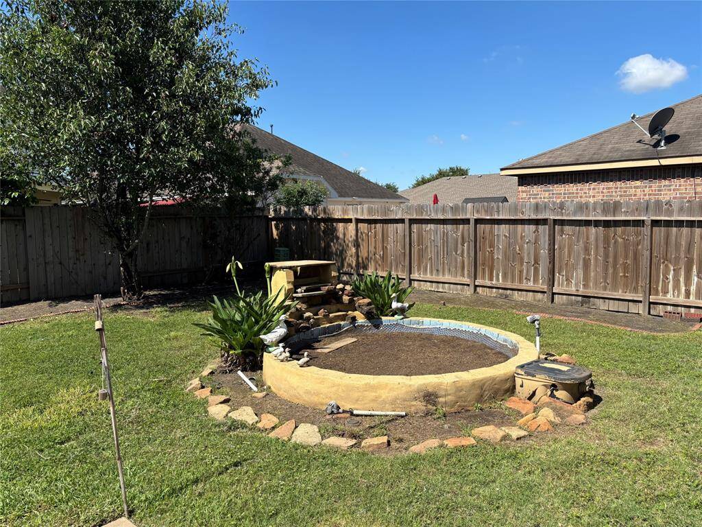 The photo features a backyard with a wooden privacy fence, a mature tree, and a small circular garden area outlined with rocks. The yard is grassy and spacious, offering potential for landscaping or outdoor activities.